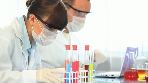 Female scientist in protective glasses and mask examines test tube with yellow liquid