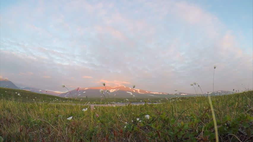 Mountains in the tundra at the Arctic circle white at night. Over the mountains quickly moving clouds. On the ground the grass grows. The mountains are illuminated by the setting sun