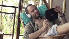 Young, happy man watching movie on tablet computer lying on hammock on terrace
