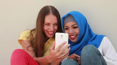 caucasian woman using phone together with asian muslim girl