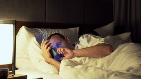 Couple in bed at night, man texting his lover on smartphone, his wife sleeping
