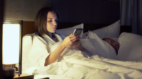 Couple in bed at night, woman texting with lover on smartphone, her husband sleeping
