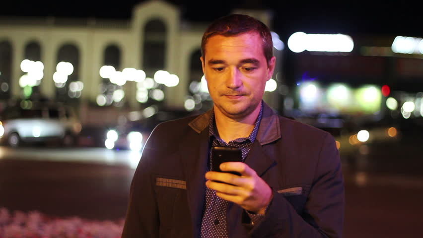 Man sms texting using app on smart phone at night in city. Handsome young business man using smartphone smiling happy wearing suit jacket outdoors.  Urban male professional