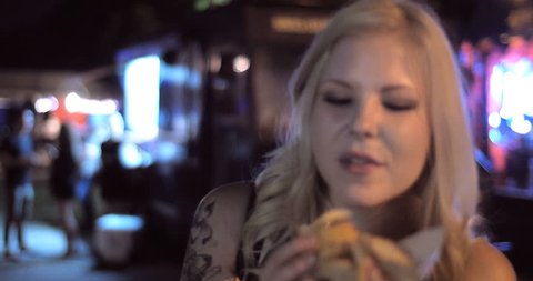 4K -A Close up shot of pretty girl eating a burger from a popular food truck in a fun urban street festival setting at night. 