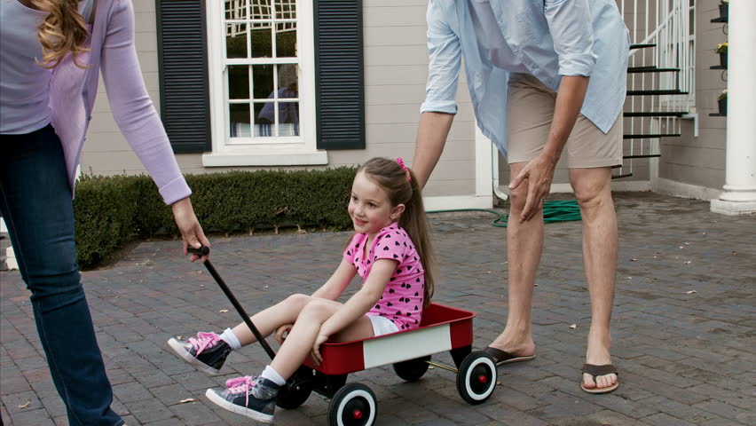 4k wide shot of father explaining to daughter how to ride for the first time on wagon as mother is just about to pull daughter in wagon down driveway. 