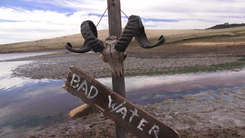 Dropping water level and a sign and skull. Bad Water sign on post at dried up water hole.