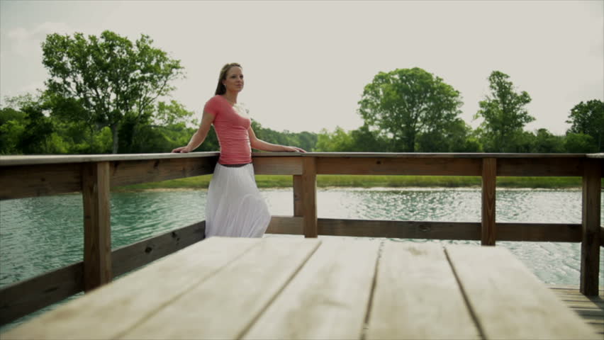 A pretty young woman wearing a long white flowing skirt standing against the railing of a pier on a very windy day. Dolly right to left
