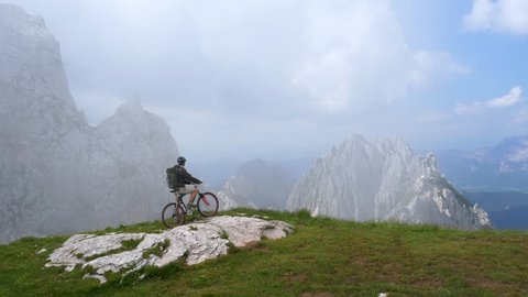 Aerial - Mountain biker looking at the view after reaching the peak. Young adult with helmet and a backpack sitting on his mountain bike and looking into the valley