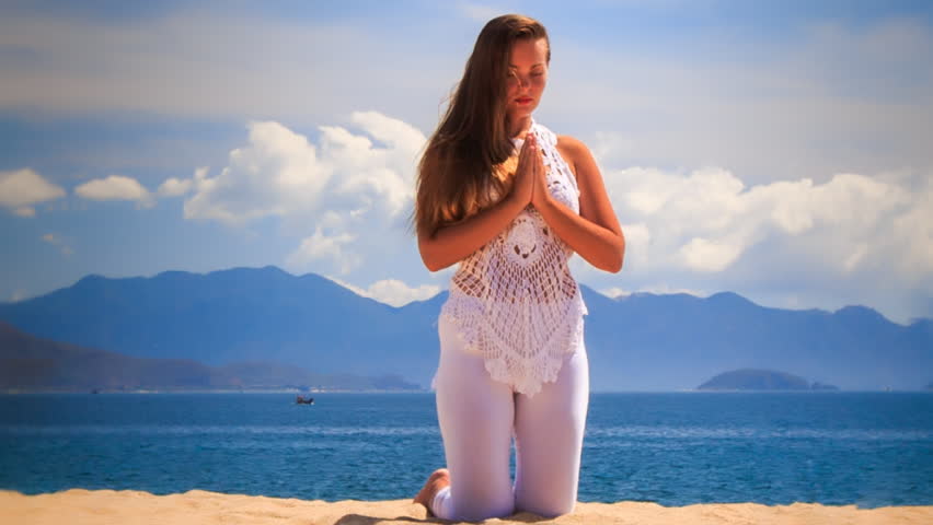 blonde girl with closed eyes in yoga asana touch hands meditates sitting on knees on beach against azure sea hills