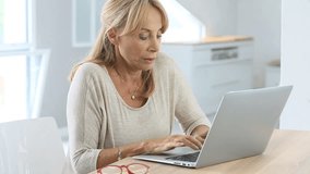 Elderly woman putting eyeglasses on while using laptop 