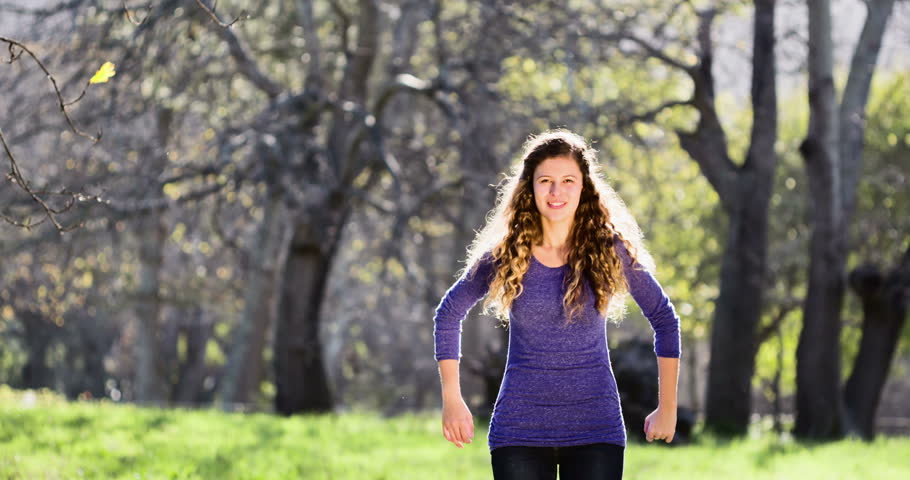 A pretty teenage girl jumps up energetically in woodland setting, hair flying and arms waving, with more enthusiastic waving after she lands. Slow motion.