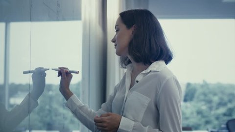 Young Female Office Worker in White Shirt is Writing on Glass Whiteboard. Shot on RED Cinema Camera in 4K (UHD).