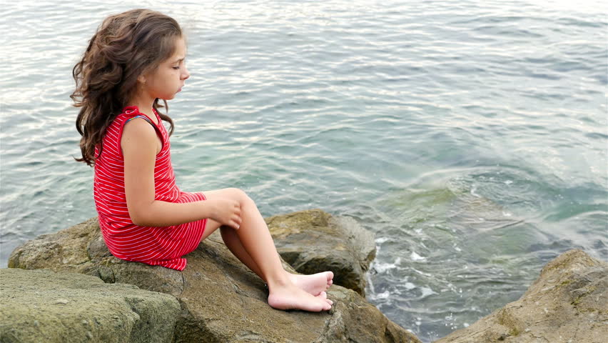 Little girl in red summer dress sitting on the rocks on the sea shore