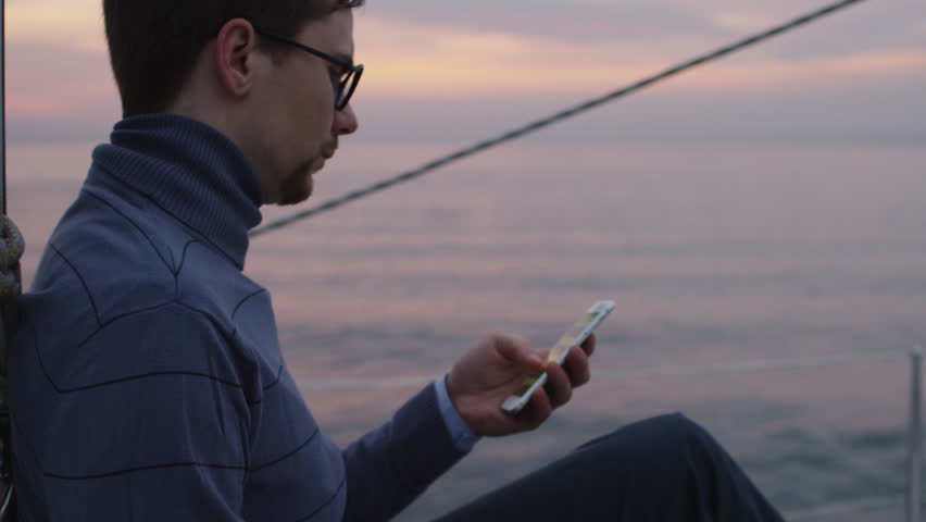 Man is looking and using phone on a sailing boat in the sea at sunset. Shot on RED Cinema Camera in 4K (UHD).