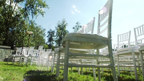 Wedding aisle decor. White wedding chairs. Outdoors wedding ceremony. Wedding set up in garden. Rows of white wooden empty chairs on lawn before wedding ceremony. Sunny day