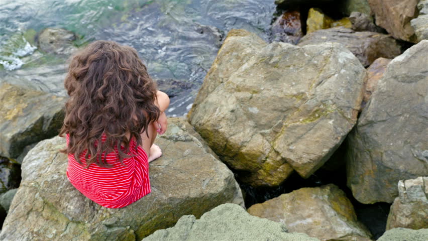 Little girl in red summer dress sitting on the rocks on the sea shore