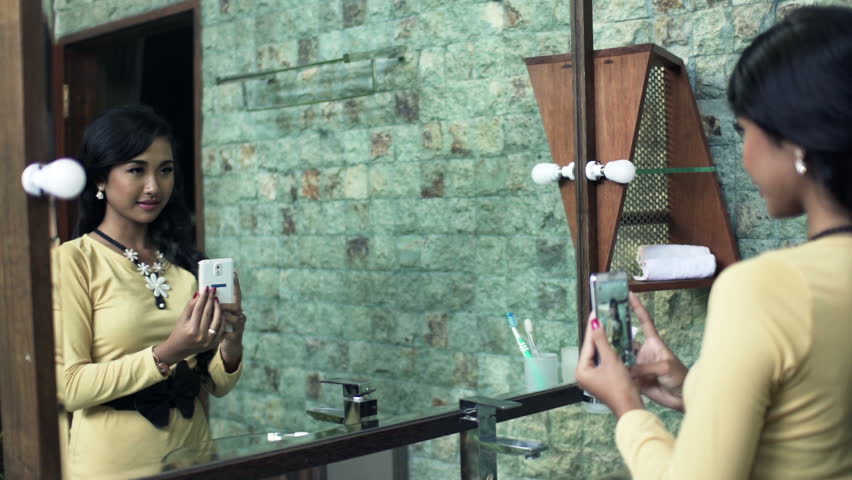 Young woman taking selfie with cellphone in front of mirror in bathroom 
