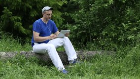 Man in dark blue t-shirt sits on a fallen tree in the forest and communicates via Tablet PC. Adult man sits on a fallen tree in the forest and uses tablet PC. Communication through video call