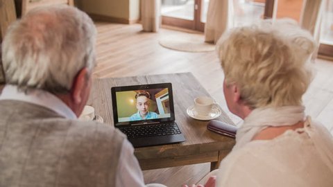 Boy waving over video call with grandparents 4K. Over shoulder view of elderly couple calling grandson with video call on laptop computer. Laptop on wooden desk. Two person sit on sofa.