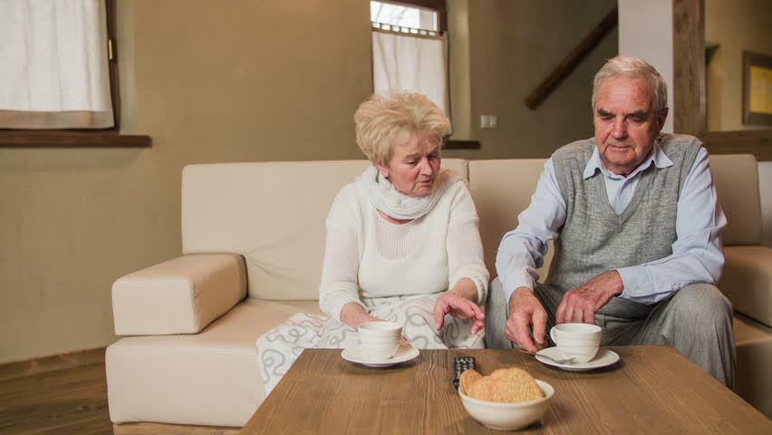 Elderly woman explaining husband about TV remote 4K. Lovey senior couple sit on sofa together and watch television with cups of tea on table. Grandma explaining husband about ownership of TV remote.