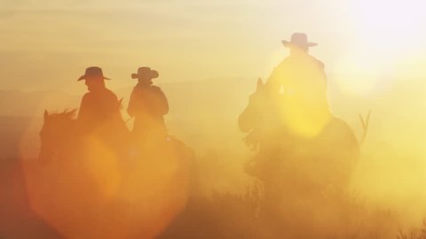 Cowboys riding into the golden sunset at dusk with mountain range background, slow motion with strong sun lens flares.