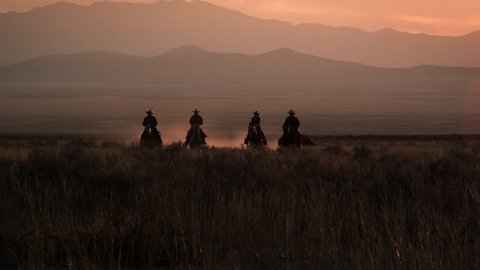 Four cowboys galloping in distance with hazy mountain range and pink sunset. Dust is visible behind horses.