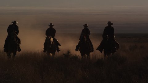 Four cowboys riding horses at dusk, slowmotion, leaving a trail of dust at sunset.