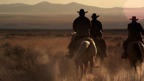 Cowboys riding off into the sunset towards mountains in the background. They leave a trail of dust behind them.