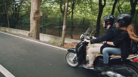 Side view of young Caucasian couple riding their scooter on beautiful country road through forest. Shot on cool autumn morning. Male shows shaka gesture to camera.