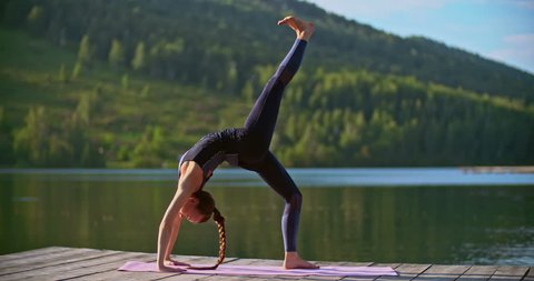 Female yoga instructor performing upward bow and one leg wheel poses on jetty over lake 