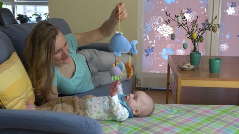 loving mother and curious baby girl play with toy on sofa. Woman and newborn daughter have fun together. Easter decoration. Tea cups and sweets on table. 4K UHD video clip.