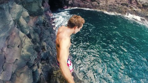 Slow Motion GOPRO POV Young Man Jumping from cliff into the ocean in Hawaii. Summer fun lifestyle. Beautiful Blue Ocean Big Sea Cave in Background. SLOW MOTION
