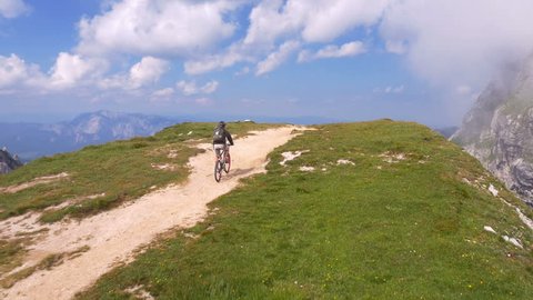 Aerial - Mountain biker arriving to the mountain top. Young adult man with a backpack enjoying sunny summer day in the mountains riding his bike to the edge of ridge