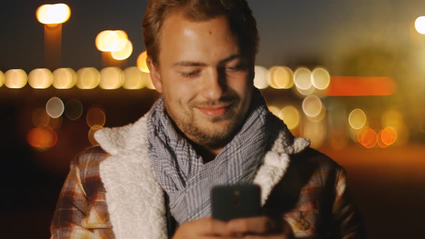 Technology - Smart handsome young man sms texting using an app on smart phone at autumn night in the city.  Using smart phone technology makes him smiling happy wearing urban hipster outfit.