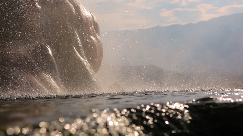 A close up shot of mist hitting the surface of a pond at the base of Fly Geyser, Nevada. The drops look like raindrops, and smooth rocks are visible in the background. Shot from water level.