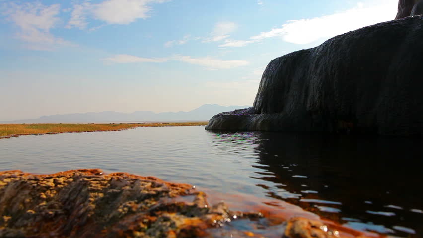 Water is flowing off of smooth rocks into a small pond below. The rocks are on the right side of the frame, in shadow. Shot at Fly Geyser in Nevada.