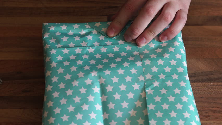 4K caucasian hands wrap starry paper around present and tape it down. A 4K shot of caucasian hands wrap blue and white starry paper around a present and taping it down with sellotape.