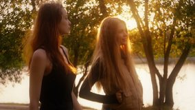 Two friends, young pretty girls, caucasians having fun outdoors in park, walking, talking, laughing, fooling around, sunset, slow motion.