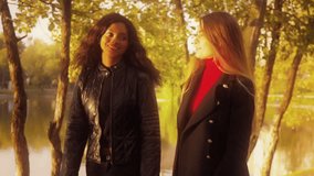 Two friends, young pretty girls, caucasian and african, having fun outdoors in park, walking, talking, laughing, fooling around, sunset, slow motion.