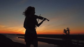 Young woman on the mountain playing the violin on sea background at sunset