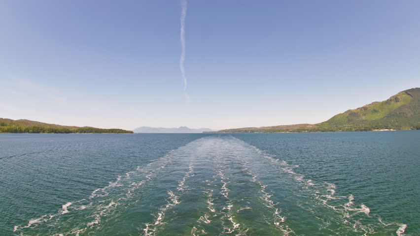 Traveling time-lapse view of the glassy ocean from behind a cruise ship, surrounded by green mountains with a clear blue sky in the background. Filmed near Ketchikan, Alaska on June 5th, 2009.