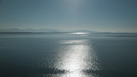 View of glistening and shimmering sea surface with hills on the horizon. Defocused glow of sun reflecting off sea water. Establishing shot, 4k. 