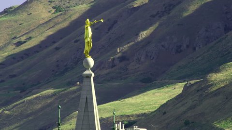 The golden-colored statue of Angel Moroni points eastward on top of the LDS Salt Lake Temple. The green and brown mountains can be seen in the background.