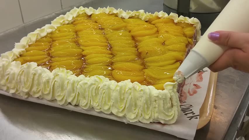 Decorating a yellow peach cake with whipped cream, at a restaurant bakery