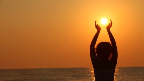 curly-headed woman standing on beach, watching sunset, holding sun in her hands