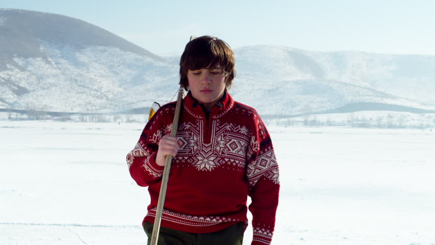 A young boy wearing a red sweater walks onto an ice covered pond holding a hockey stick and staring forward with anticipation.