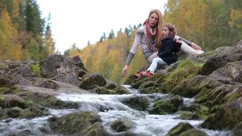 A young mother and her cute daughter sitting near the forest stream and play with water.