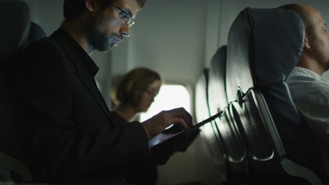 Young man is using a tablet on an airplane and a woman is reading in the background next to a window. Shot on RED Cinema Camera in 4K (UHD).