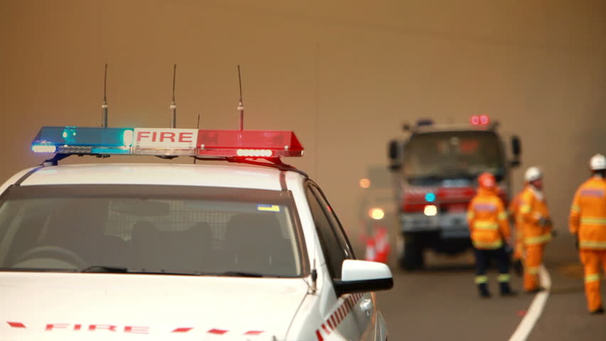 The fire car is the point of focus in this shot. Out of focus firemen work next to the fire truck in the background. They are working hard to contain the bush fire that is out of shot, on camera right