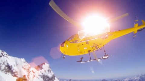 Action sportlers were dropped by a helicopter at the top of the mountains. The sun is shining brightly in the blue sky. There is a mountain range in the background covered in snow.